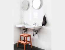 Zwei weiße Waschbecken mit runden Spiegeln darüber an einer gefliesten Wand, daneben hängt ein gestreiftes Handtuch. Ein orangefarbener Tritt steht vor den Waschbecken auf einem dunklen Boden.