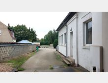 Ein gepflasterter Hof zwischen einem weißen Gebäude mit mehreren Fenstern und einer grauen Mauer aus Betonsteinen. Im Hintergrund sind Bäume sowie geparkte Autos zu sehen.