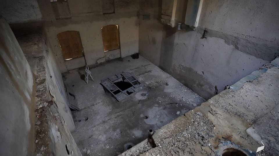 Blick in einen großen, leerstehenden Betonraum mit hohen Wänden und staubigem Boden; in der Mitte befindet sich eine rechteckige Öffnung mit Metallrahmen. An der Stirnseite sind zwei hohe Fenster mit Holzbrettern vernagelt, daneben sind raue Betonflächen und eine abgesenkte Nische erkennbar.
