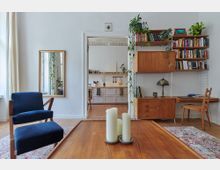 Wohnraum mit hellen Wänden und Holzboden, eingerichtet mit einem blauen Sessel, einem niedrigen Holztisch mit Kerzen, einem Regal mit Büchern und Pflanzen sowie einer Durchgangstür zur Küche. Die Küche im Hintergrund hat weiße Schränke, offene Regale und eine helle, minimalistische Gestaltung.