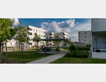 Moderne Wohnanlage mit mehreren weißen, mehrstöckigen Gebäuden, umgeben von Grünflächen und jungen Bäumen. Im Vordergrund befinden sich ein überdachter Fahrradabstellplatz und ein gepflasterter Gehweg.