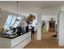 Eine moderne, helle Wohnküche mit offener Gestaltung. Zu sehen sind ein großer Küchentresen mit schwarzer Oberfläche, darüber zwei Kronleuchter, ein Esstisch vor schrägen Dachfenstern und ein angrenzendes Wohnzimmer mit weißen Wänden, einer freigelegten Ziegelwand und Holzböden.