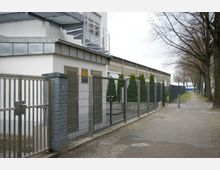 Ein moderner Zaun mit durchsichtigen Metallgittern umgibt ein Gebäude mit weißer Fassade und Warnschildern an Metalltüren. Daneben verläuft ein Bürgersteig mit Pflastersteinen, gesäumt von Bäumen und Grünflächen.