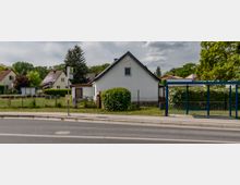 Bushaltestelle an einer Straße mit blauem Unterstand, daneben ein weißes Einfamilienhaus mit spitzem Dach und ein eingezäuntes Grundstück. Im Hintergrund sind weitere Häuser und Bäume zu sehen.