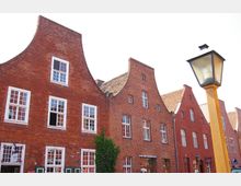 Ziegelrote Häuserfassaden mit geschwungenen Giebeln in einer historischen Straße; Fenster mit weißen Rahmen und eine gelbe Straßenlaterne im Vordergrund.
