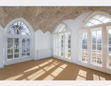 Heller Raum mit hohen Spitzbogenfenstern, die nach außen auf eine Terrasse mit Holzmöbeln führen. Die dekorativ bemalte Gewölbedecke zeigt florale Muster, und der Boden ist mit einem beigefarbenen Teppich ausgelegt.