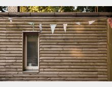 Holzwand eines Gebäudes mit horizontalen Brettern, einem schmalen Fenster mit milchiger Glasscheibe und einer Wimpelkette aus bunten Stoffdreiecken, die darüber hängt. Im Hintergrund sind Bäume und Blätter zu erkennen.