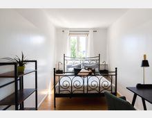 Helles Schlafzimmer mit einem schwarzen Metallbett im Mittelpunkt, vor einem Fenster mit weißen Vorhängen. Links ein Regal mit einer Topfpflanze, rechts ein Schreibtisch mit einer Lampe, auf dem Boden Parkettboden.