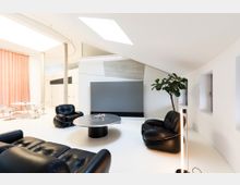 Modernes Wohnzimmer mit weißen Wänden und schräger Decke, ausgestattet mit schwarzen Ledersesseln, einem runden Tisch und einer langen Stehleuchte. Im Hintergrund befinden sich eine rosa Gardine und ein Esstisch mit transparenten Stühlen, sowie eine große Zimmerpflanze und mehrere kleine Fenster.