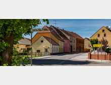 Straßenszene in einem kleinen Dorf mit gelben und rosa Wohngebäuden, teils mit Ziegeldächern. Im Vordergrund befinden sich ein Baum, ein Straßenschild mit der Aufschrift "Karl-Liebknecht-Allee" und Wegweiser zu nahegelegenen Orten.