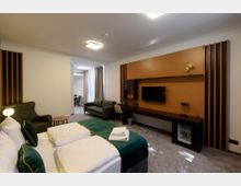 Ein modernes Hotelzimmer mit einem großen Bett im Vordergrund, grünen Akzenten und weißen Kissen. Im Hintergrund befinden sich ein Schreibtisch mit Flachbildfernseher, ein Sofa, ein Sessel, ein kleiner Tisch sowie bodenlange Vorhänge vor großen Fenstern.