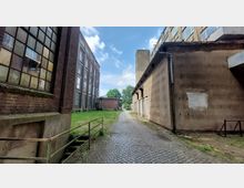 Hinterhof eines alten Fabrikgeländes mit Pflasterweg, flankiert von einem Backsteingebäude mit hohen Fenstern auf der linken Seite und einem verputzten Gebäude auf der rechten Seite. Im Hintergrund sind eine kleine Wiese, Bäume und ein weiterer Gebäudeteil sichtbar.