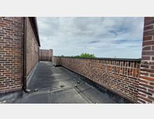 Flachdach eines Backsteingebäudes mit umlaufender Backsteinbrüstung und schwarzer Dachabdichtung. Im Hintergrund sind Bäume und ein bewölkter Himmel zu sehen.