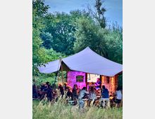 Eine Outdoor-Szene mit einer Gruppe von Menschen, die unter einem weißen Pavillon sitzen, umgeben von hohen Gräsern und Bäumen. Im Hintergrund ist eine beleuchtete Backsteinwand mit einem kleinen Fenster zu sehen.