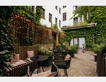 Innenhof mit kleinem Cafébereich, gepflastertem Boden und mehreren runden Tischen mit geflochtenen Stühlen und Sitzkissen. Die hellen Gebäudefassaden und Holzelemente sind stark mit Efeu und Pflanzen bewachsen; am Rand stehen Pflanzkübel und ein Fahrrad.