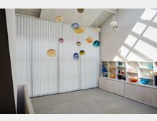Heller, minimalistischer Raum mit hängenden, farbigen Glaslampen und einem Regal an der rechten Wand, gefüllt mit dekorativen Glasobjekten in verschiedenen Farben. Im Hintergrund befindet sich ein großer Vorhang, der weiches Tageslicht durchlässt.