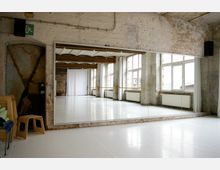 Ein Tanzstudio mit einer Wand aus großen Spiegeln, weißen Wänden mit sichtbaren Backsteinen und großen Fenstern, die viel Licht hereinlassen. Der Raum hat helle Bodenbeläge, Heizkörper unter den Fenstern und teilweise freiliegende Metall- und Holzelemente.