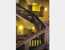 Treppenhaus in einem historischen Gebäude mit massiven Steinbalustraden, gelb gestrichenen Wänden und Bögen, die von Säulen getragen werden. An den Wänden sind runde Wandlampen befestigt, die den Raum beleuchten.