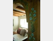 Ein Holzpfosten mit geschnitztem und bemaltem Blumendekor in Blau- und Grüntönen, im Hintergrund ein gut beleuchtetes Wohninterieur mit Holzbalkendecke und einer Sitzecke mit Kissen.