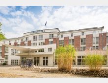 Fassade eines modernen Hotels mit dem Schriftzug „Inselhotel“ über dem Eingang, mit weißen und roten Klinkerelementen. Im Vordergrund befinden sich eine Überdachung, ein Fahrradständer und Ziergräser auf einem gepflasterten Platz.