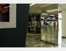 Innenraum einer modernen Galerie mit glänzendem Marmorboden, Glaswänden und Spiegeln, die den Raum optisch vergrößern. An den Wänden sind gerahmte Kunstwerke ausgestellt, und im Hintergrund befinden sich beleuchtete Deckenpaneele.