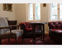 Wohnzimmer im klassischen Stil mit einer roten Ledercouch, einem gepolsterten Sessel mit floralem Muster und einem Holztisch. Im Hintergrund sind weiße Fenster mit Blick auf Bäume sowie eine antike Kommode und ein gerahmtes Gemälde an der Wand zu sehen.