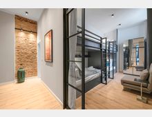 Moderner Schlafsaal im Hostelstil mit mehreren schwarzen Etagenbetten und Vorhängen, hellen Holzböden und weißen Wänden. Links ist eine freiliegende Ziegelwand mit Wandlampen, am Ende ein Spiegel und rechts ein kleines Sitzmöbel zu sehen.