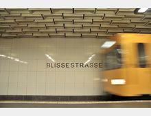 U-Bahn-Station "Blissestraße" mit gefliester Wand, auf der der Stationsname steht, und einem vorbeifahrenden gelben Zug im Vordergrund. Die Decke zeigt ein geometrisches Muster aus rechteckigen Vertiefungen.