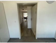 Blick durch eine geöffnete weiße Tür in einen Raum mit hellem Laminatboden; im Hintergrund ein Essbereich mit einem Tisch und Stühlen sowie ein Fenster mit Blick nach draußen. Der Vordergrund zeigt einen Boden mit grauen Fliesen.