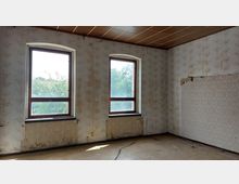 Ein leerer Raum mit zwei großen, rechteckigen Fenstern, durch die Tageslicht einfällt und Bäume draußen sichtbar sind. Die Wände sind mit verblasster und teilweise abblätternder Tapete bedeckt, die einen floralen oder ornamentalen Stil aufweist, und der Boden zeigt Abnutzungs- und Beschädigungsspuren.