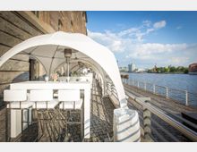 Außenbereich eines Restaurants oder Cafés direkt am Wasser, mit weiß überdachten Sitzbereichen mit hohen Tischen und Stühlen auf Kopfsteinpflaster neben einer Uferpromenade. Im Hintergrund ist ein Fluss mit Gebäuden und einer Stadtlandschaft sichtbar.