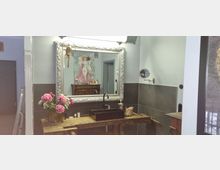 Badezimmer mit rustikalem Waschbecken auf einer hölzernen Arbeitsplatte, einem schwarzen Wasserhahn und einer großen, verzierten Wandspiegel. Im Hintergrund ist ein Gemälde sowie ein antikes Möbelstück zu sehen, dazu Dekorationen wie ein Blumenstrauß und Pflegeprodukte.