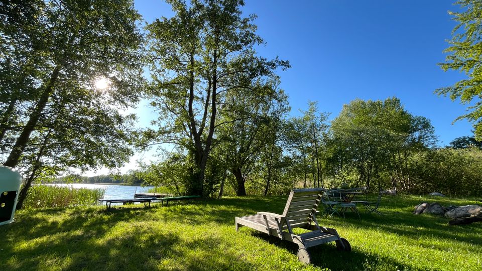 Wiese am Ufer eines Sees mit hohen Laubbäumen und Blick auf einen kleinen Steg im Wasser. Im Vordergrund stehen eine hölzerne Gartenliege und ein Metallgartentisch mit Stühlen, umgeben von Sträuchern und einzelnen Steinen unter blauem Himmel.