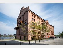 Backsteingebäude mit fünf Etagen im industriellen Stil, teilweise mit kleinen Balkonen und begrünten Bereichen umgeben. Im Vordergrund führt eine gepflasterte Straße, während im Hintergrund ein Fluss und moderne Gebäude zu sehen sind.