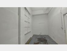 Schmaler, weiß gestrichener Raum mit abgenutztem Bodenbelag, links ein großes Fenster mit weißem Rahmen, hinten eine schmale Tür, teilweise sichtbare Kabel an den Wänden.