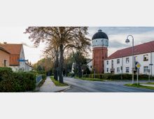 Straßenszene mit einem Gehweg, einer Baumreihe und Häusern mit roten Ziegeldächern; im Hintergrund ein roter Backstein-Wasserturm mit weißer Kuppel. Die Umgebung wirkt ländlich und ruhig.