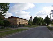 Straßenszene in einem Dorf mit einem gelben Backsteingebäude links und einem Fachwerkturm mit rotem Spitzdach im Hintergrund. Auf der rechten Seite stehen Bäume entlang einer gepflasterten Straße, die von Bürgersteigen gesäumt ist.