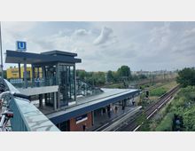 Außenansicht einer U-Bahn- und S-Bahn-Station in einer urbanen Umgebung, mit einem überdachten Eingangsbereich, einer gläsernen Aufzugsanlage und Treppen. Im Hintergrund sind Bahngleise, Grünflächen und Gebäudestrukturen zu sehen.