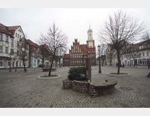 Weitläufiger gepflasterter Marktplatz mit kahlen Bäumen, umgeben von historischen Gebäuden mit roten Dächern. Im Hintergrund steht ein markantes rotes Backsteingebäude mit Giebeln und einem Uhrenturm.