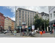 Straßenecke mit mehrstöckigem Hotelgebäude im modernen Stil und umliegenden älteren Gebäuden mit bunten Fassaden. Im Vordergrund befinden sich eine Uhr, Fahrradständer, Werbetafeln und Straßenverkehr, während im Hintergrund Bäume sichtbar sind.