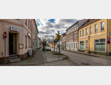 Straßenszene in einer kleinen Stadt mit gepflastertem Gehweg, mehrfarbigen historischen Gebäuden und einem Baum in der Straßenmitte. Rechts sind gelbe und weiße Fassaden mit großen Fenstern, links ein älteres Gebäude mit einem Ladenschild, das "Jonis Oase" anzeigt.
