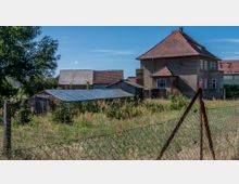 Ein ländliches Grundstück mit einem zweistöckigen Wohnhaus mit braunem Verputz und einem roten Ziegeldach. Neben dem Haus befinden sich Nebengebäude mit Wellblechdächern, umgeben von einer Wiese mit hohen Gräsern und Büschen, eingezäunt durch einen Maschendrahtzaun.