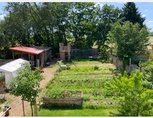 Ein Garten mit mehreren Hochbeeten, Gemüsebeeten und jungen Bäumen, umgeben von Bäumen und Holzzäunen. Im Hintergrund sind eine einfache Holzhütte mit rotem Dach sowie ein offenes Zelt zu sehen.