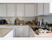 Moderne Küche mit weißen Hochglanzschränken, hellen Holz-Arbeitsplatten und integriertem Waschbecken. Auf den Arbeitsflächen stehen diverse Küchengeräte, Vorratsgläser und ein Tablett mit Gewürzen und kleinen Behältern.