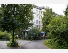 Wohnhaus im Altbaustil mit heller Fassade, umgeben von üppiger Vegetation und Bäumen. Im Vordergrund befindet sich ein kleiner gepflasterter Weg, daneben ein Café mit Schirmen und Fahrrädern.