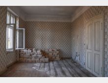 Ein leerer Raum mit verblasster, gemusterter Tapete, einem Holzboden und einer geöffneten Fensterflügel auf der linken Seite. An der Wand steht ein Stapel heller Ziegelsteine, und rechts befindet sich eine weiße Holztür mit dekorativen Paneelen.
