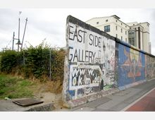 Ein Abschnitt der bemalten Berliner Mauer, bekannt als East Side Gallery, mit Graffiti und Kunstwerken auf der Oberfläche. Im Hintergrund sind ein modernes Gebäude und eine bewachsene Hecke mit Metallzaun sichtbar.