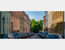 Eine von Häuserreihen gesäumte Kopfsteinpflasterstraße mit parkenden Autos auf beiden Seiten; im Hintergrund sind grüne Bäume zu sehen, die einen Übergang zu einer parkähnlichen Umgebung andeuten. Die Fassaden der Gebäuden sind in verschiedenen Pastellfarben gestrichen und zeigen dekorative Gesimse und Fensterumrahmungen.