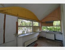 Ein kleiner Innenraum mit gefliesten Böden, halbhohen Fenstern und einem gelben, gebogenen Holzpaneel an der Decke. Die Fenster zeigen Blick auf Bäume, während Heizkörper und ein einfacher Arbeitstisch unter den Fenstern sichtbar sind.