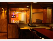 Ein traditioneller Kegelbahnbereich mit Holzvertäfelung, Schreibtisch und Stühlen im Vordergrund sowie Blick durch ein Fenster auf die beleuchtete Kegelbahn im Hintergrund.